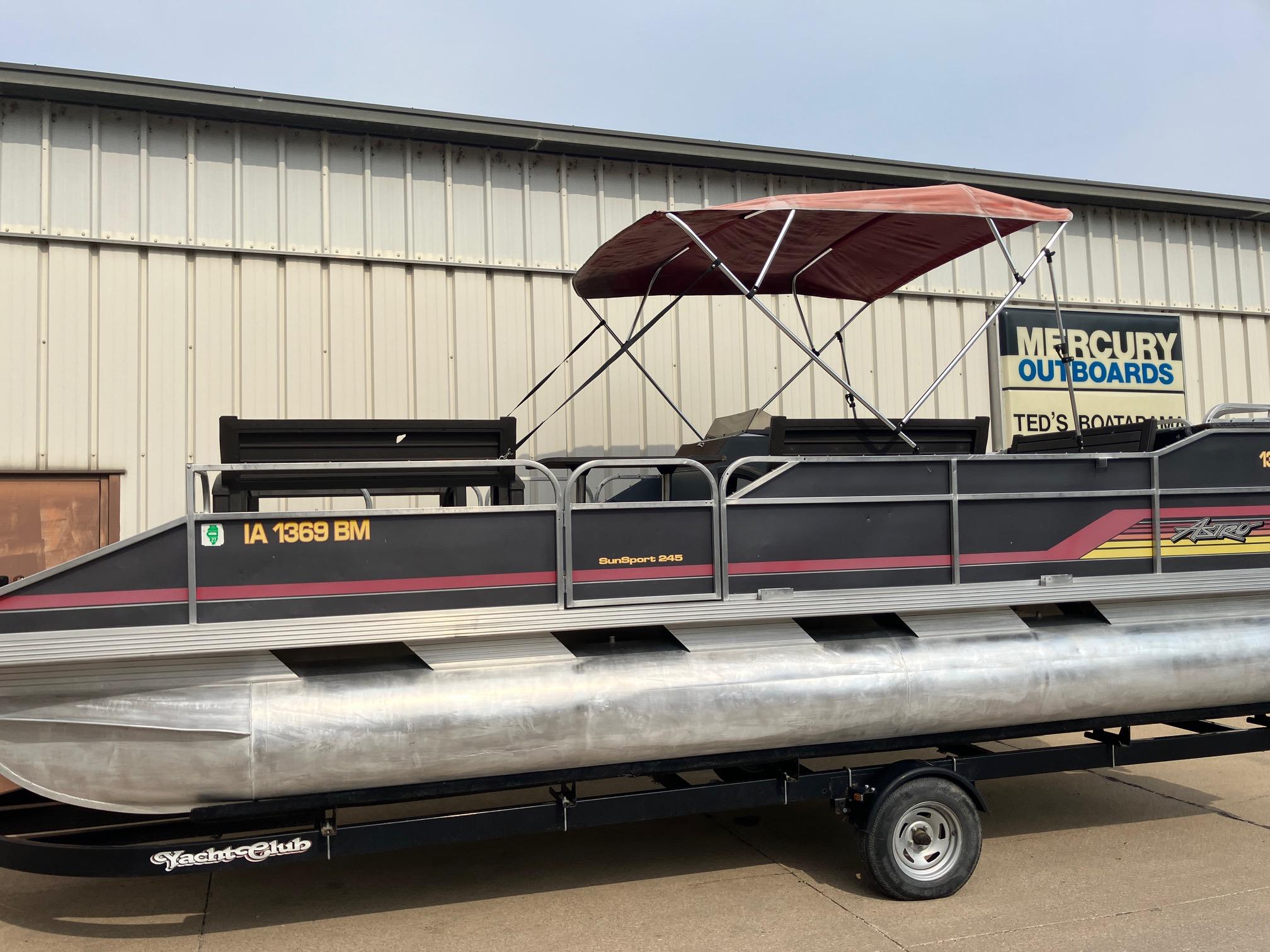 1990 Astro 24' Pontoon boat with canopy on trailer, parked outside Mercury Outboards shop.