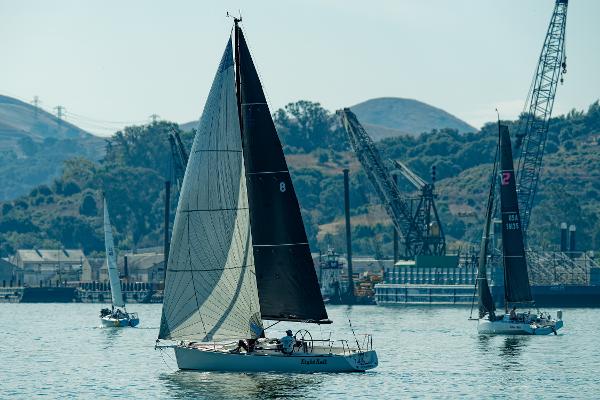 Sailboats on calm water, featuring a 2005 J Boats J/100, with industrial cranes in the background.