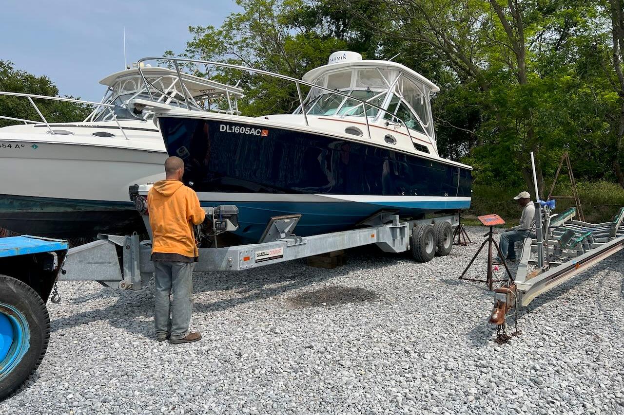 2006 Back Cove 26 boat on trailer, two people nearby, surrounded by trees and gravel.