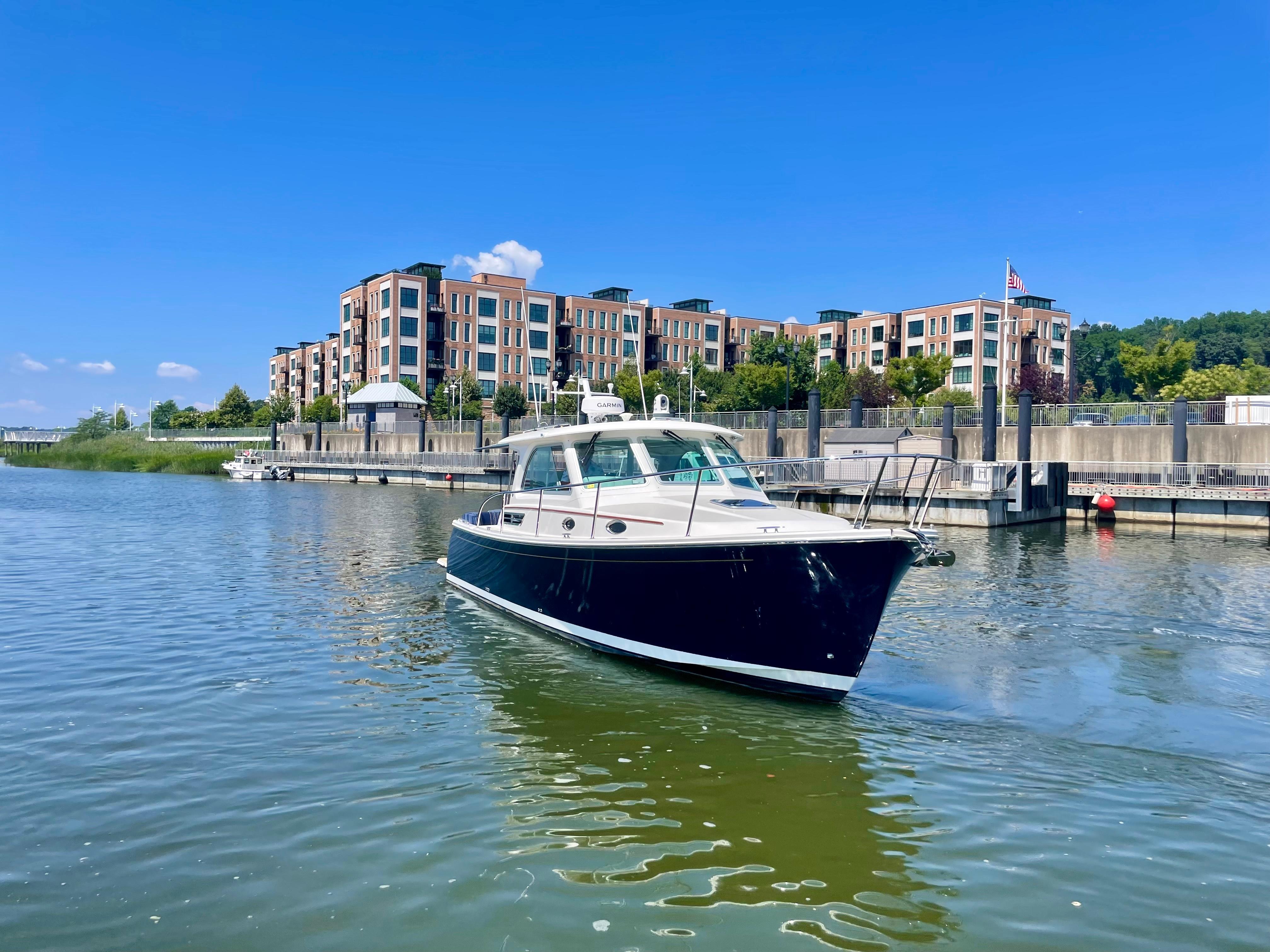 2023 Back Cove 34O boat on a calm river near modern waterfront buildings.