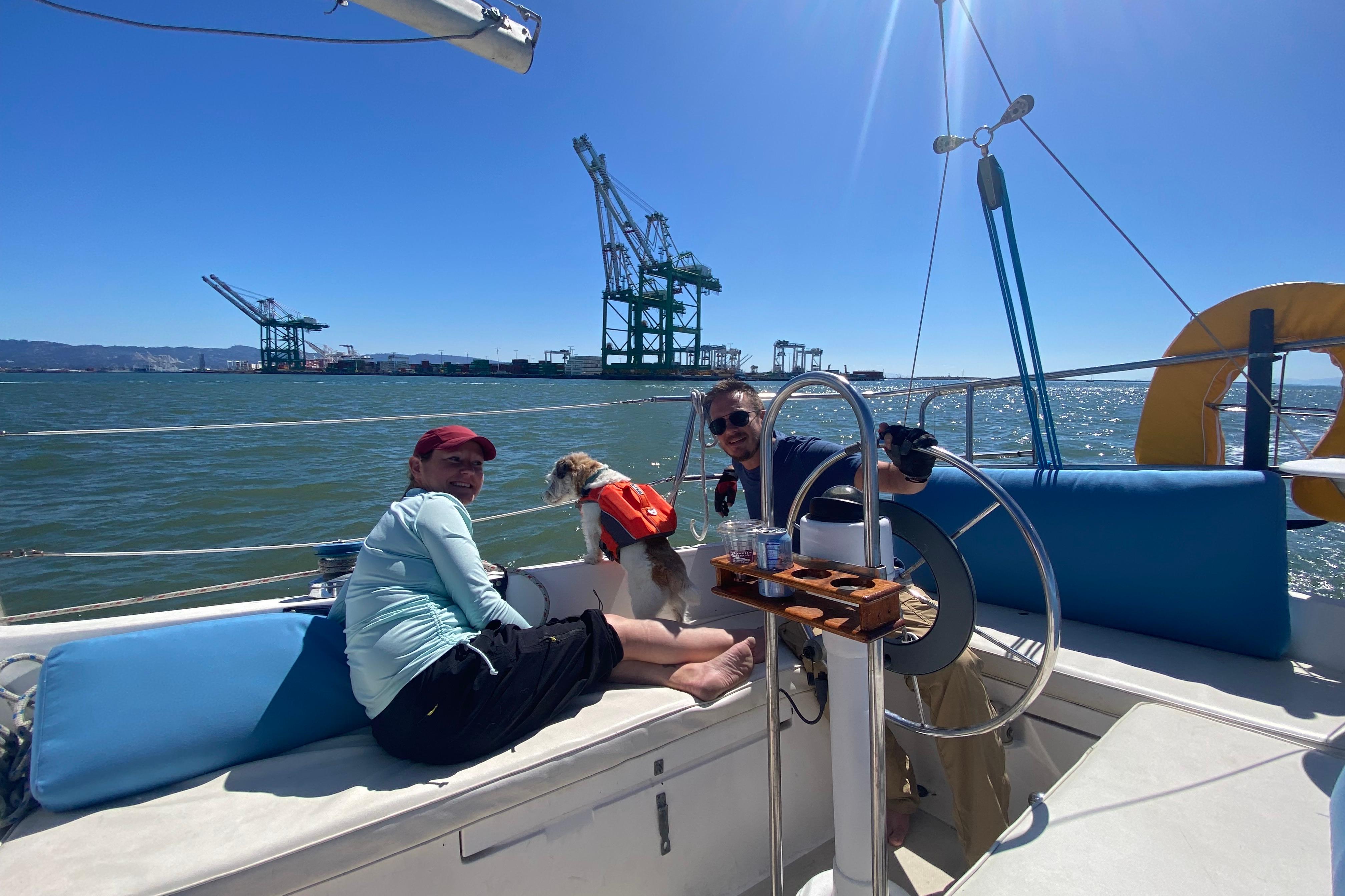 Two people and a dog on a 1980 Catalina 30 sailboat, with cranes in the background.