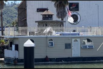 Floating home docked by a marina, featuring a rooftop deck and nautical design elements.