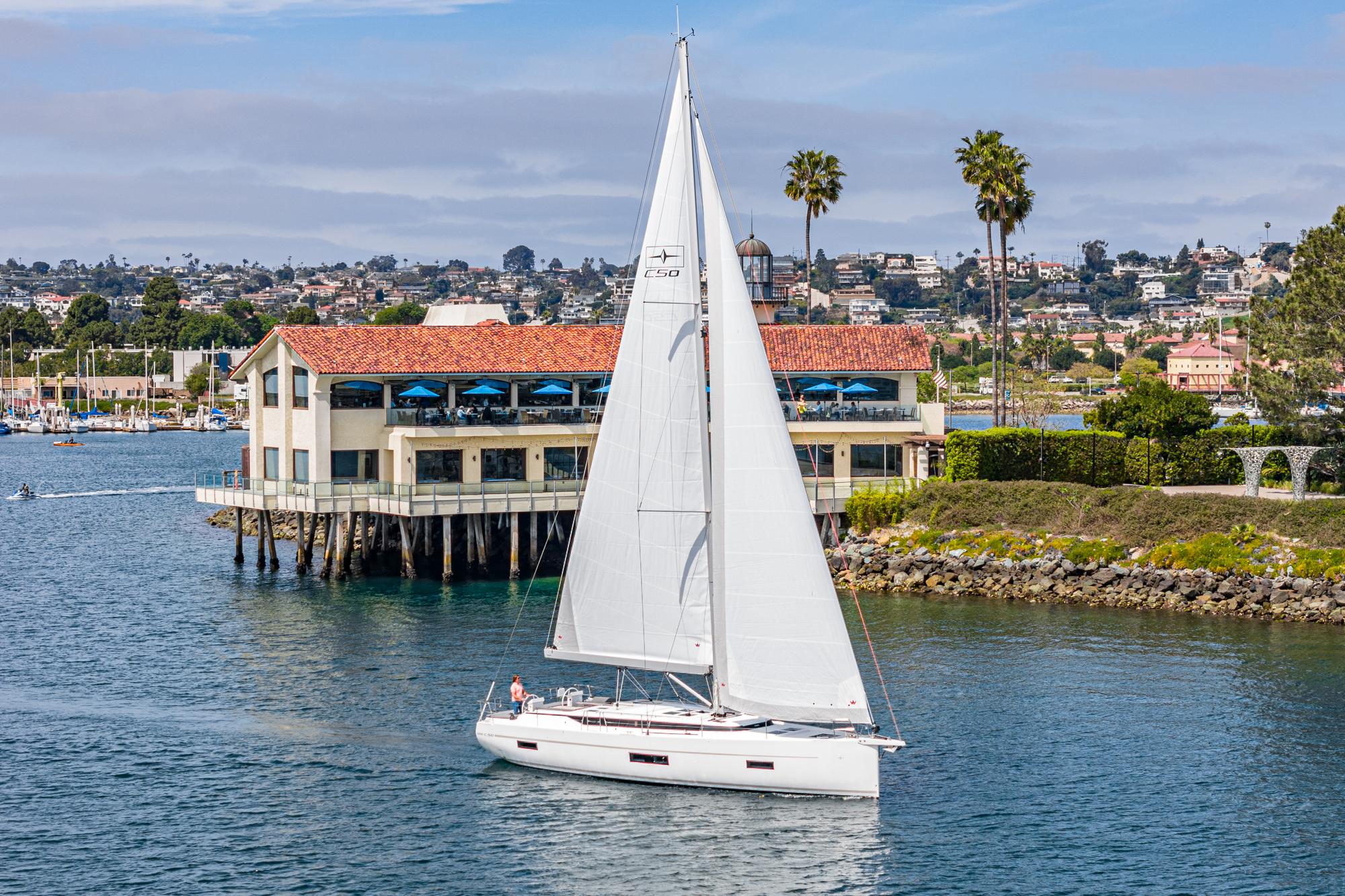Bavaria-C50 2024-This could be yours CA-US-2024 Bavaria C50 sailboat cruising near a coastal restaurant with palm trees.-9654457 | Thumbnail