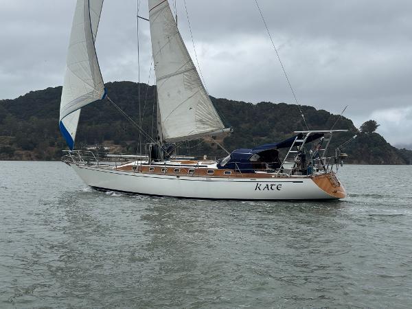 1983 Tayana 52 Aft Cockpit sailboat on water, sails up, overcast sky, scenic background.