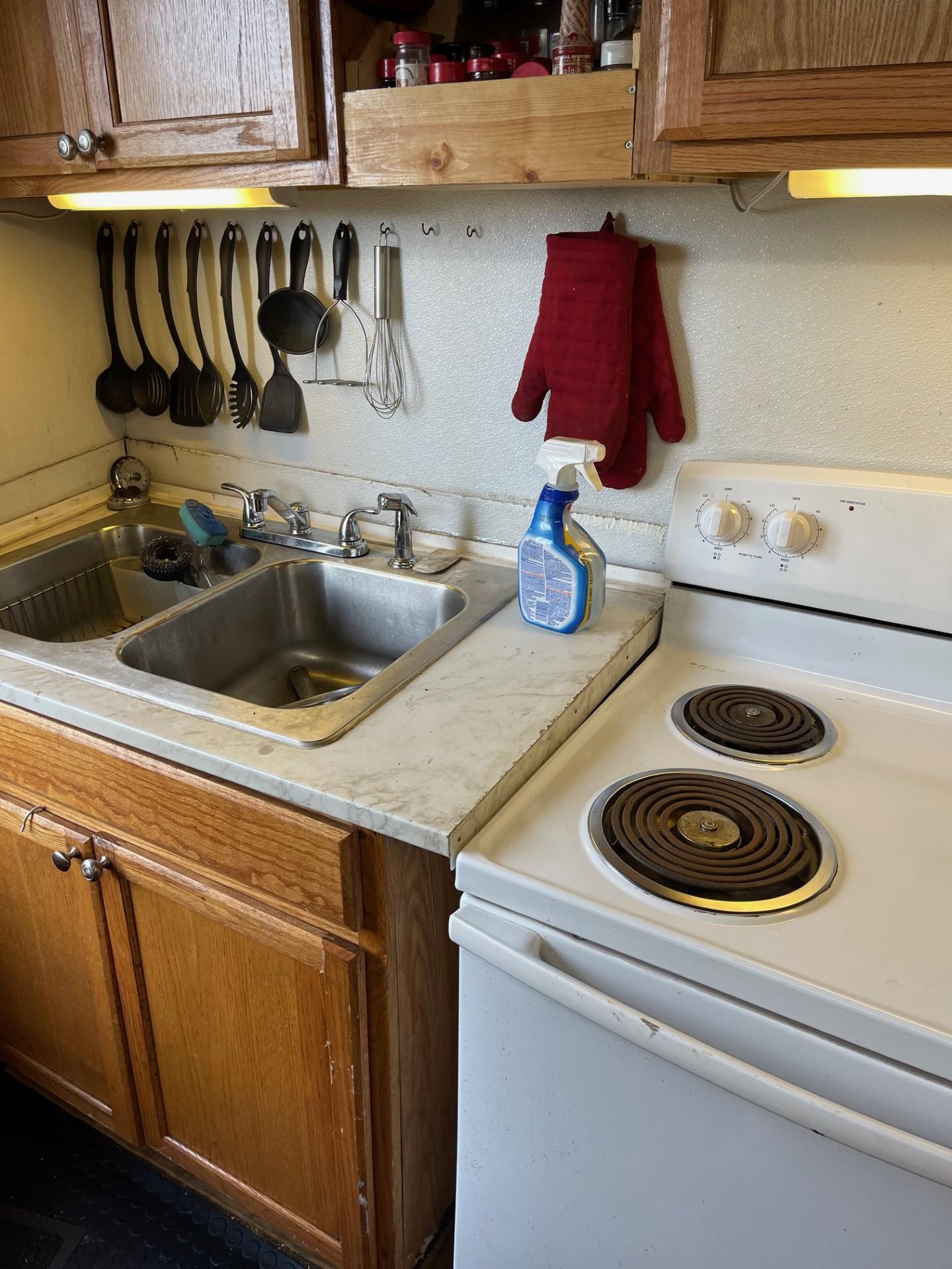 Commercial-Model Bow Tug 1961 -FL-US-Compact kitchen with wooden cabinets, dual sink, electric stove, and cleaning supplies.-9787034 | Thumbnail