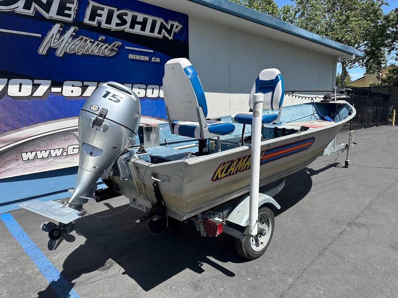 1998 Klamath 14 DLX boat with outboard motor on trailer, parked outside marine shop.