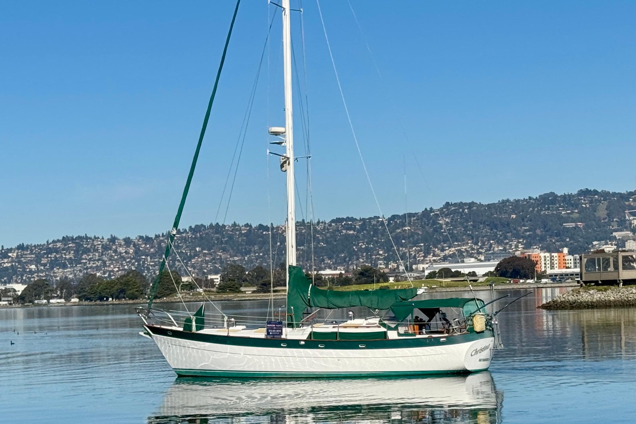 Sailboat "Christina" by Hans Christian, 1988, on calm water with scenic hillside backdrop.