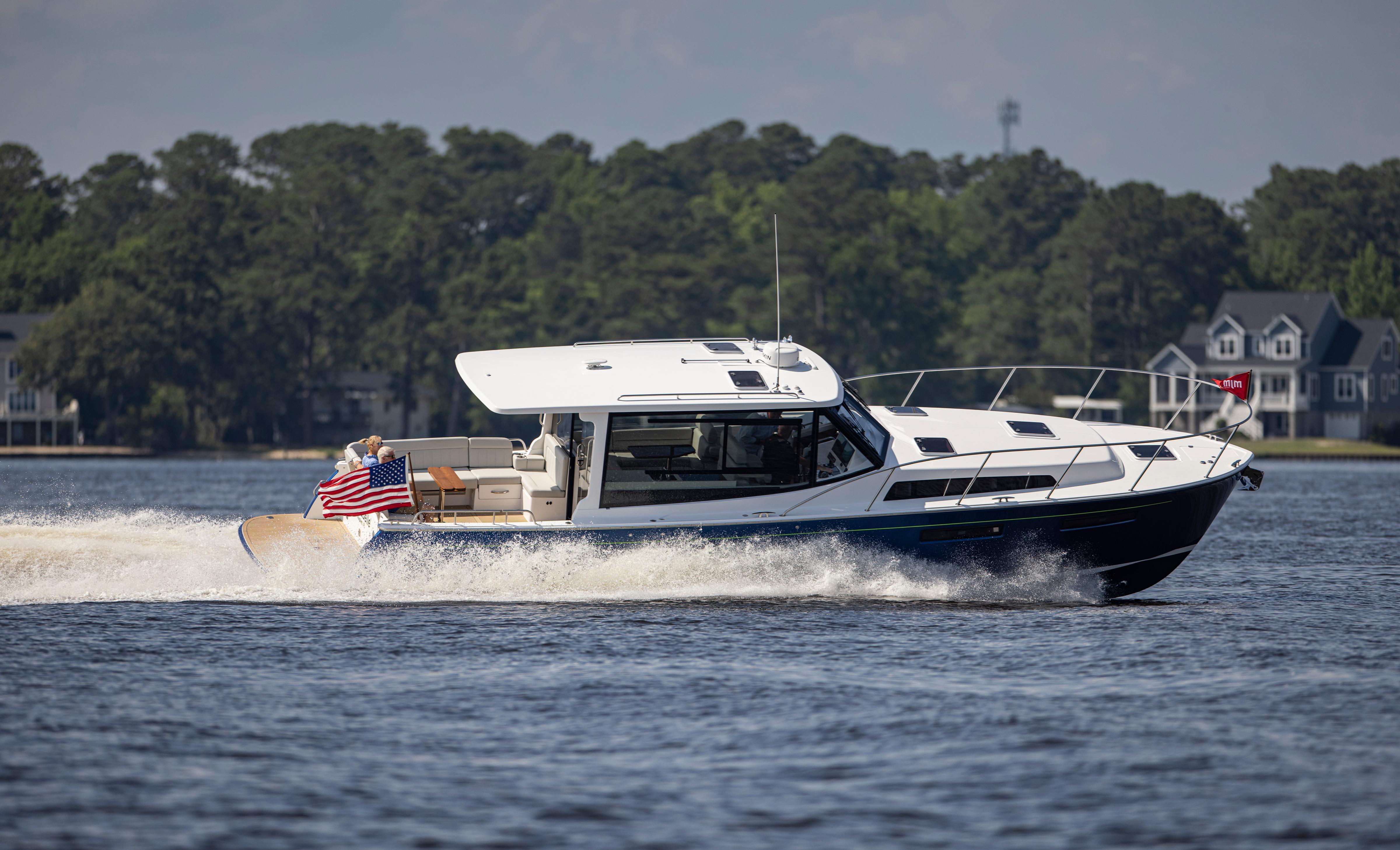 2025 MJM 42 yacht cruising on a lake with American flag, surrounded by trees and houses.