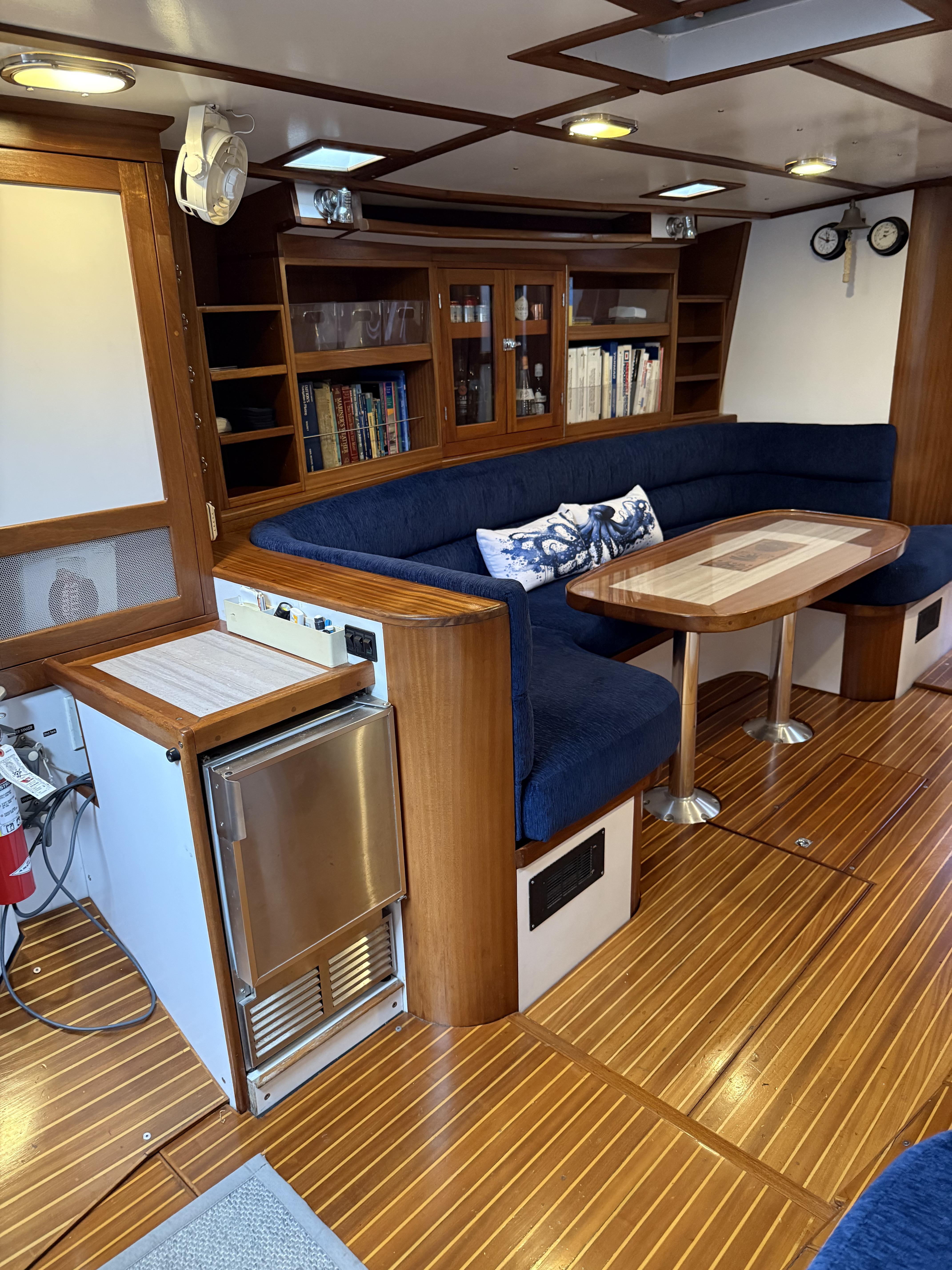 Luxurious 1976 Frers Custom yacht interior with blue seating, wooden table, and built-in bookshelves.