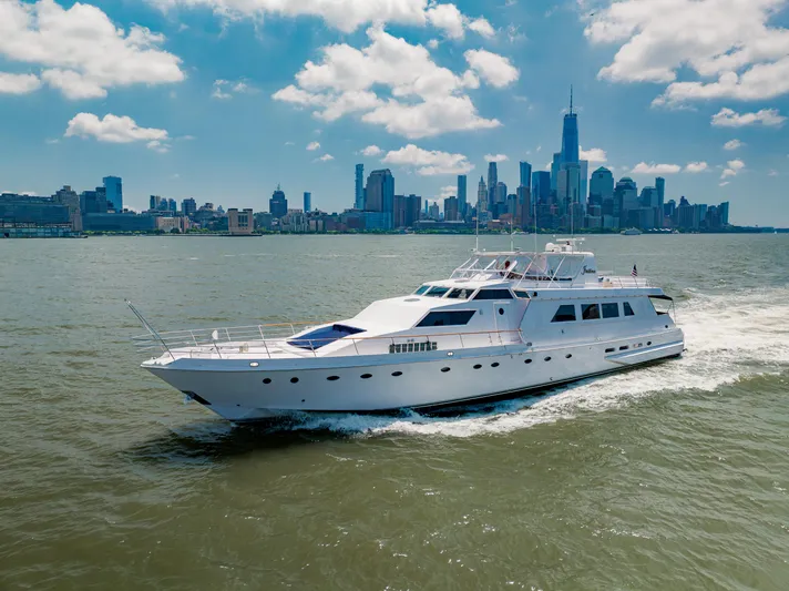 Justine Yacht Photos Pics Luxurious 1987 Guy Couach 97 yacht cruising near a city skyline under a blue sky.