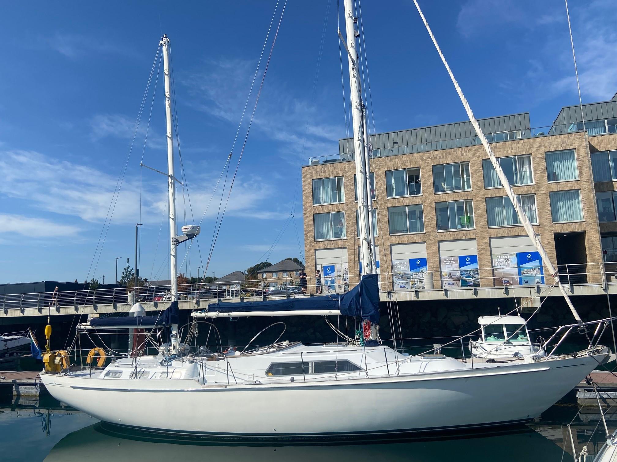 1978 Nicholson 48 Ketch sailboat docked in marina, clear sky, modern building backdrop.