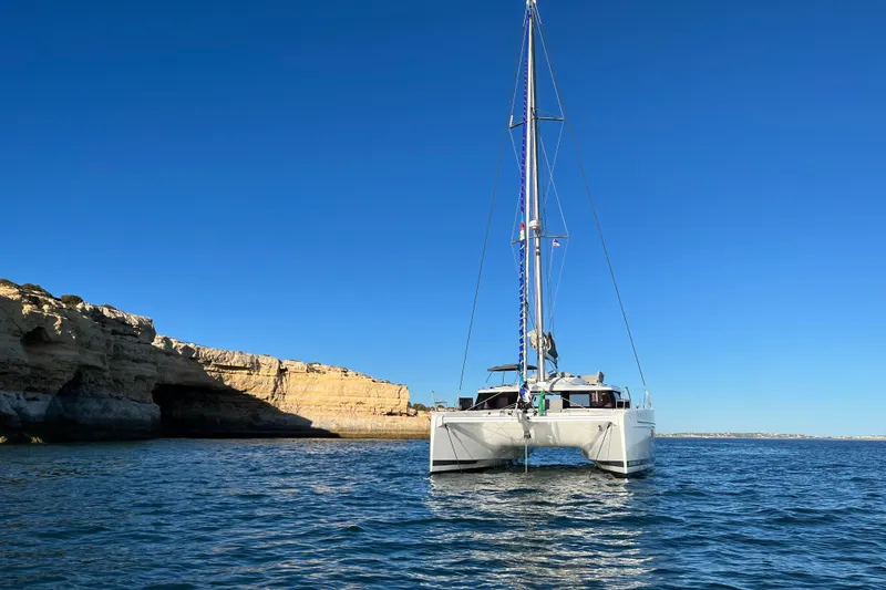Free To Be Yacht Photos Pics 2022 Fountaine Pajot Elba 45 Catamaran sailing near rocky coastline under clear blue sky.