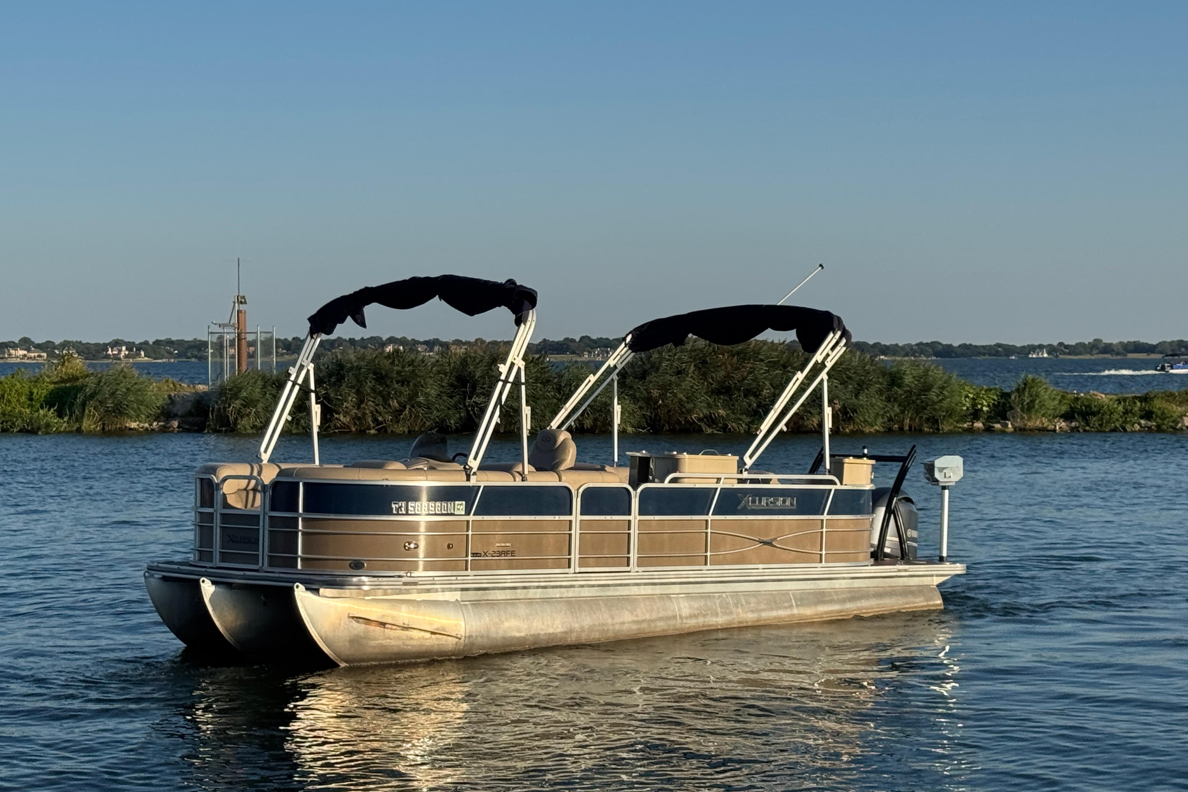 2016 Xcursion 23RFE pontoon boat on a serene lake with lush greenery.
