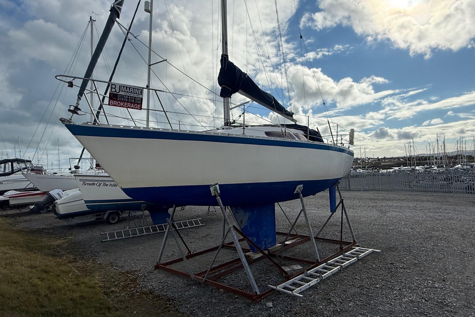 1984 Trapper T30 sailboat on stands, brokerage sign, cloudy sky background.