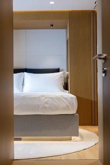 Mini K2 Yacht Photos Pics Modern bedroom with white bedding, wooden accents, and soft lighting, viewed through an open door.