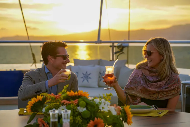 Aresteas Yacht Photos Pics Couple enjoying drinks on a yacht at sunset, surrounded by flowers.
