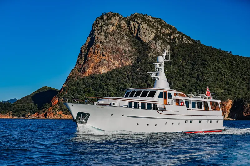 Kapal Yacht Photos Pics Classic 1971 Feadship yacht cruising near a rocky coastline under a clear blue sky.