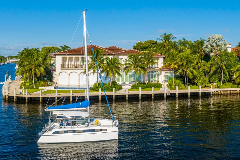  Yacht Photos Pics Luxury catamaran Smart Cat S280 sailing near waterfront mansion, surrounded by palm trees and clear blue sky.
