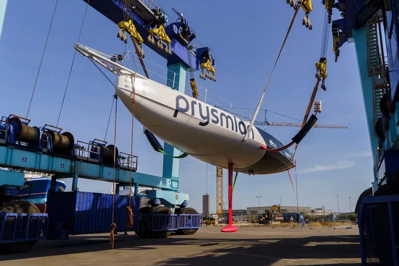 Prysmian Yacht Photos Pics Imoca 60 sailboat by Multiplast, 2015 model, being lifted by cranes at a shipyard.
