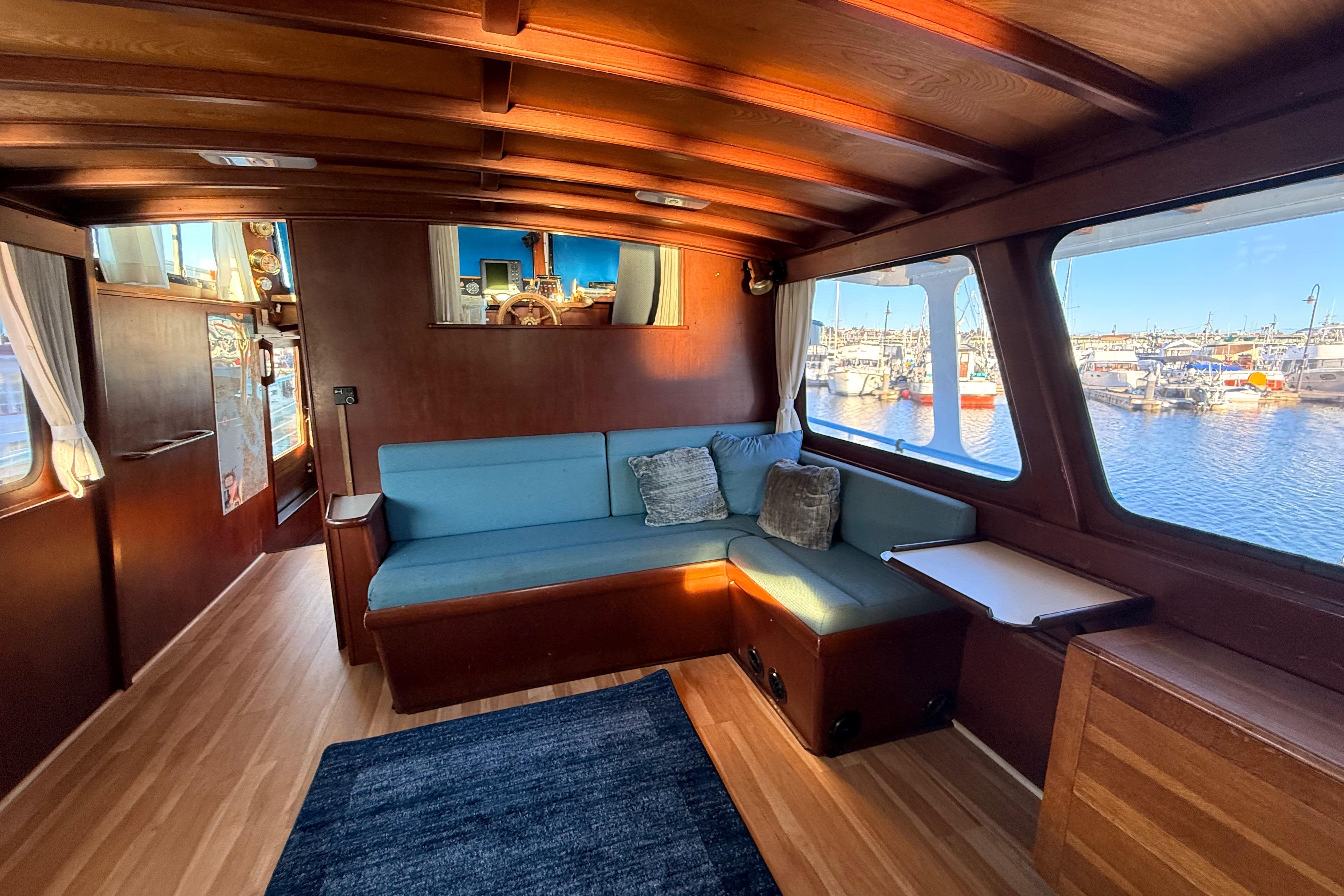Interior of a 1965 Custom Trawler with wooden paneling and blue seating, overlooking a marina.