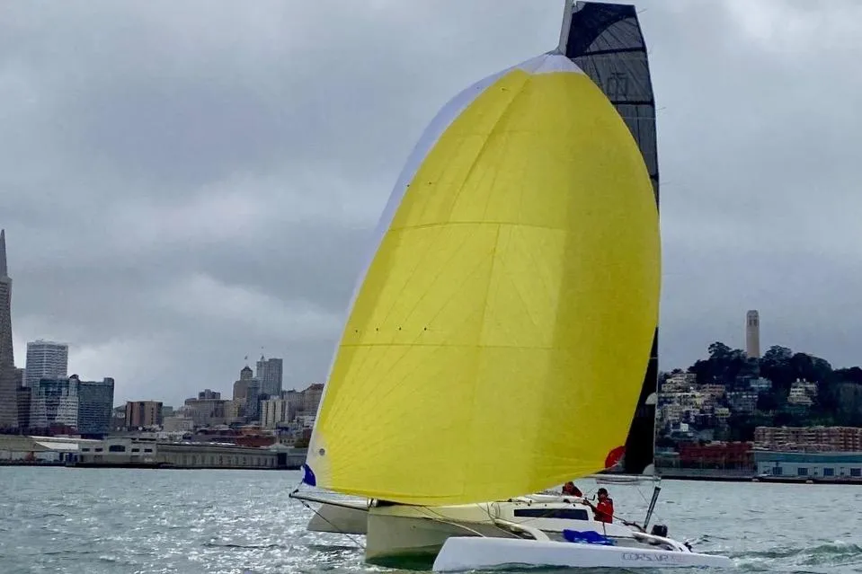 Sailboat Corsair 760 Dash 2018 with yellow sail on a cloudy day near city skyline.