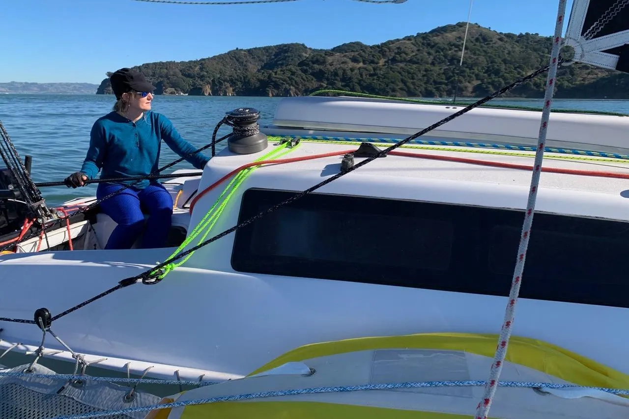 Person sailing a 2018 Corsair 760 Dash on calm waters near a forested coastline.