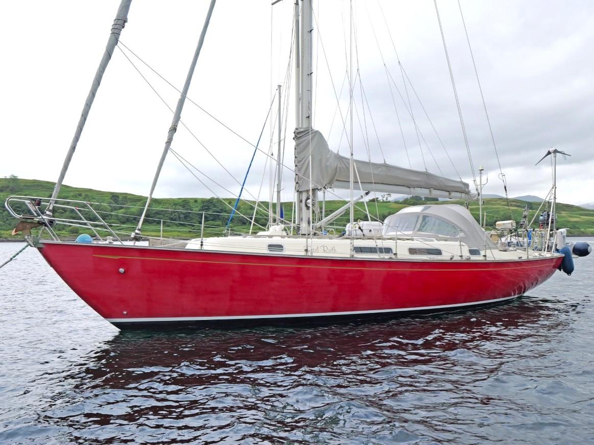 Red 1999 Rustler 42 sailboat on calm water, with green hills in the background.