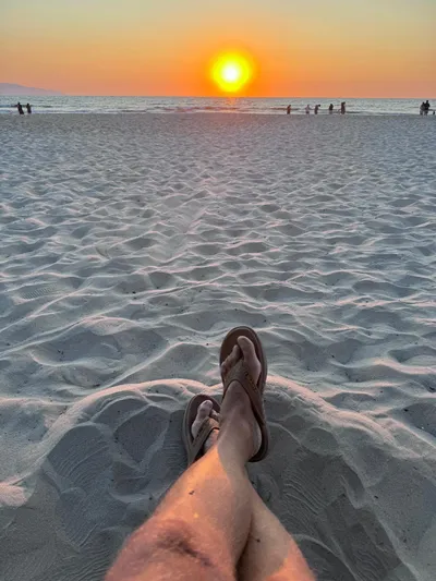 La Ultima Chica Yacht Photos Pics Person relaxing on sandy beach at sunset, wearing flip-flops, with distant ocean view.