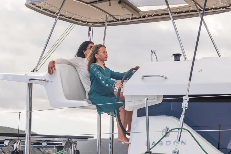  Yacht Photos Pics Young girl steering 2026 Lagoon 38 catamaran, with adult seated nearby, under cloudy sky.