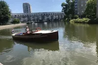 Vintage 1926 custom motorboat cruising on a calm urban river with modern buildings in the background.