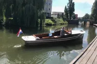 Custom 1926 motorboat cruising on a canal with a Dutch flag, surrounded by greenery and buildings.