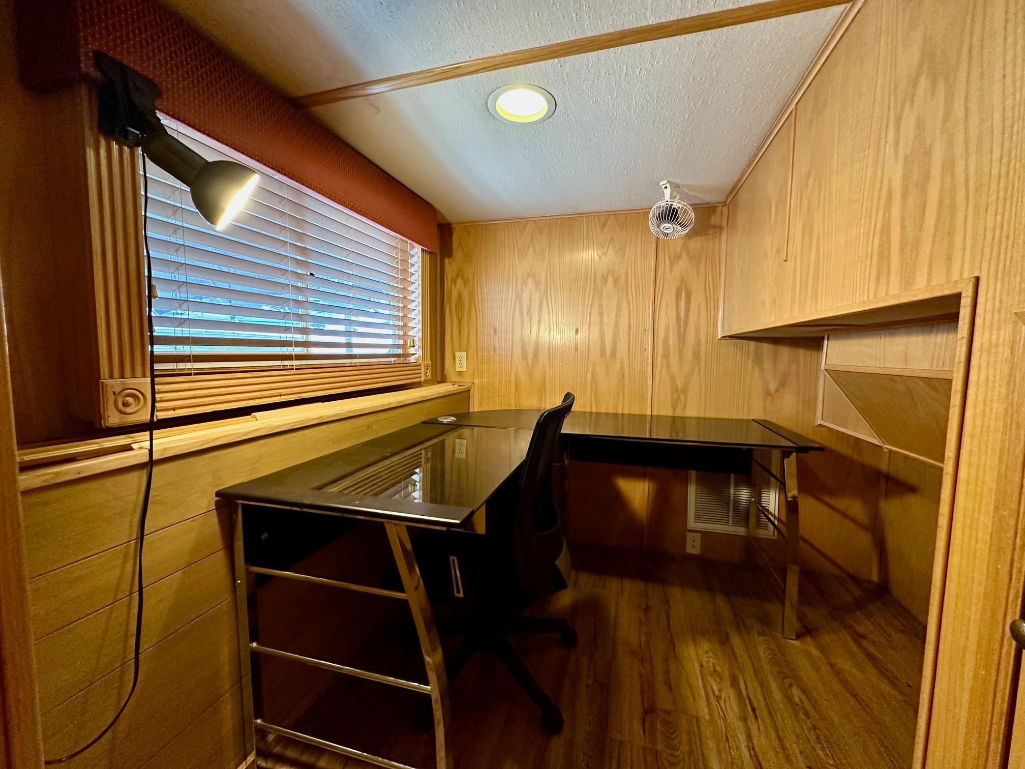 Small wooden office space with desk, chair, and window blinds in a 2004 Lakeview 16x68 model.