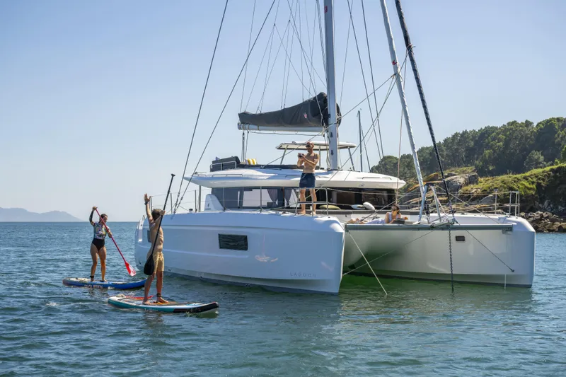  Yacht Photos Pics 2025 Lagoon 43 catamaran with people paddleboarding nearby in a scenic coastal setting.