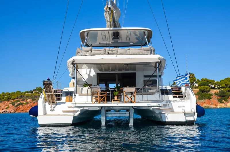 Esperance Yacht Photos Pics 2024 Lagoon 55 catamaran on water, rear view, with Greek flag and scenic coastline.