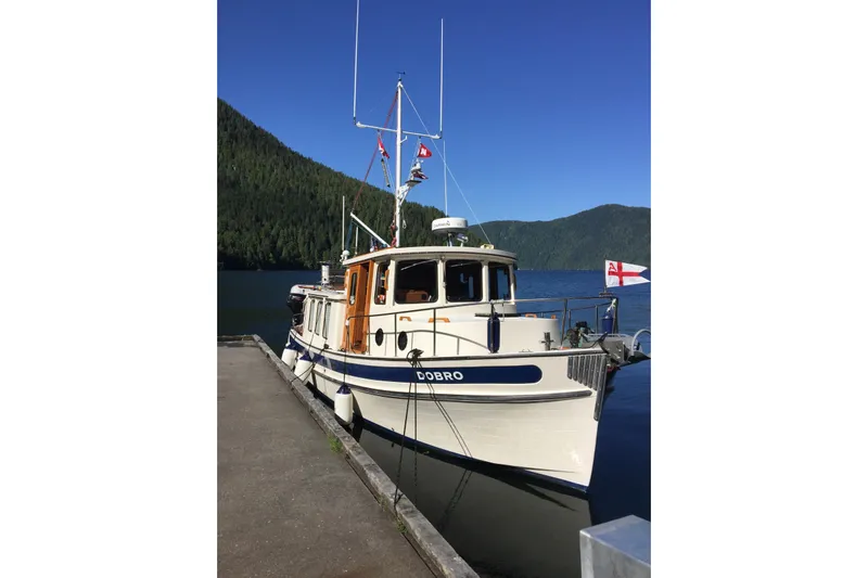 Dobro Yacht Photos Pics 1990 Nordic Tug 32 docked by scenic forested lake under clear blue sky.