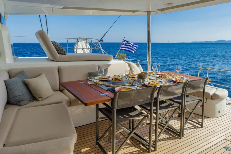  Yacht Photos Pics Luxurious 2023 Lagoon 55 yacht deck with dining setup, overlooking serene blue sea and Greek flag.