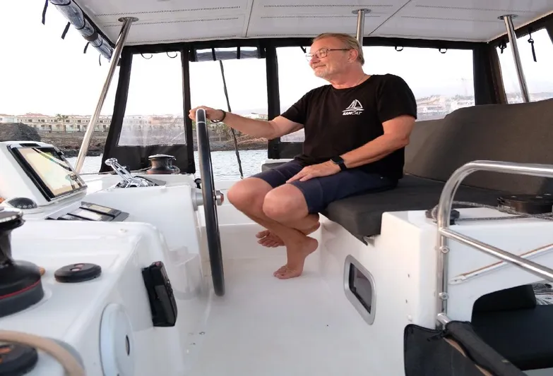 Samcat Yacht Photos Pics Man steering a 2020 Lagoon 46 catamaran, seated at the helm, coastal background.