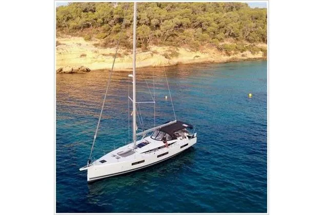  Yacht Photos Pics 2021 Jeanneau 60 sailboat anchored in clear blue water near a wooded shoreline.