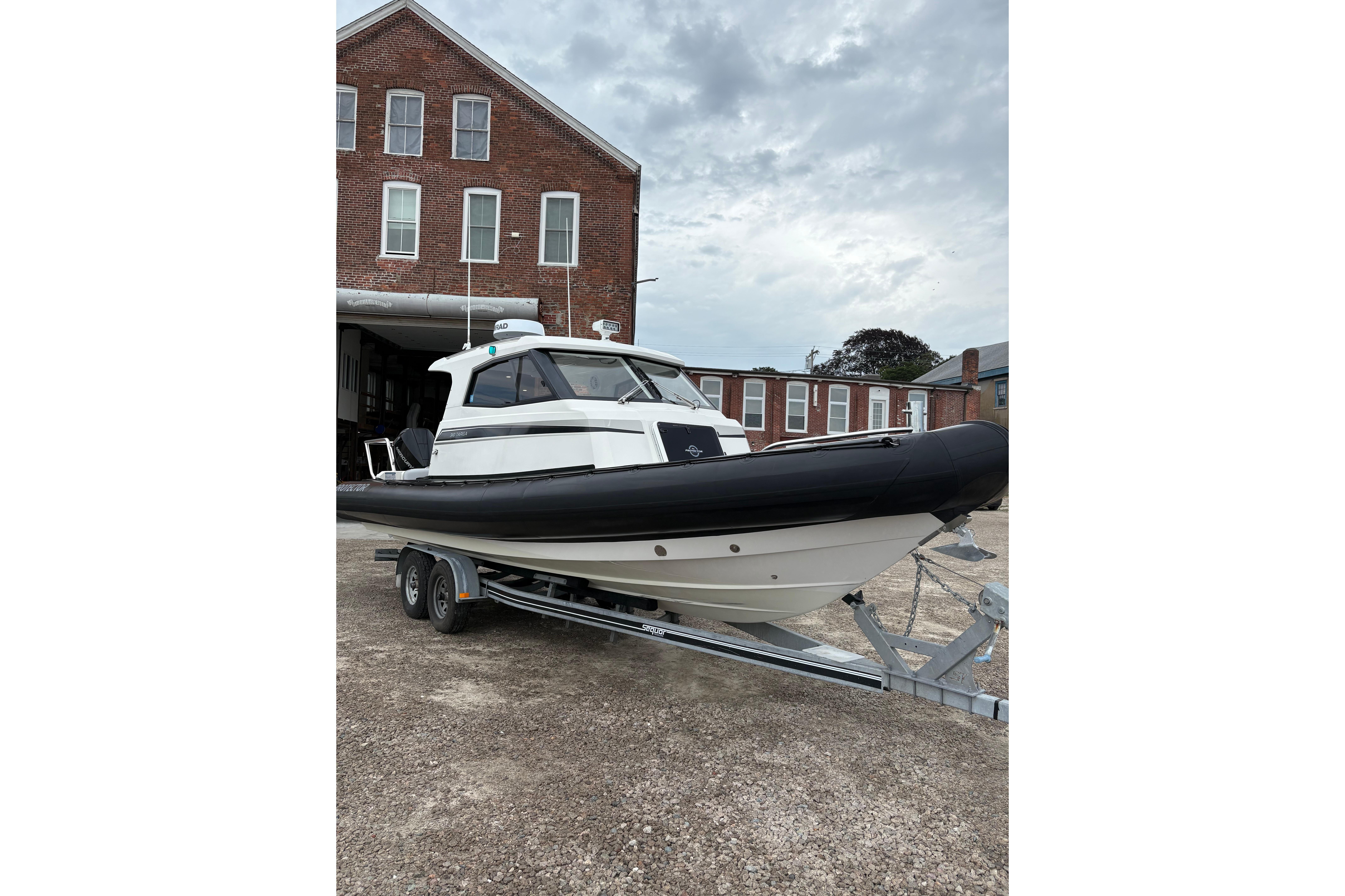 2020 Protector 310 Targa boat on trailer, parked near brick building under cloudy sky.