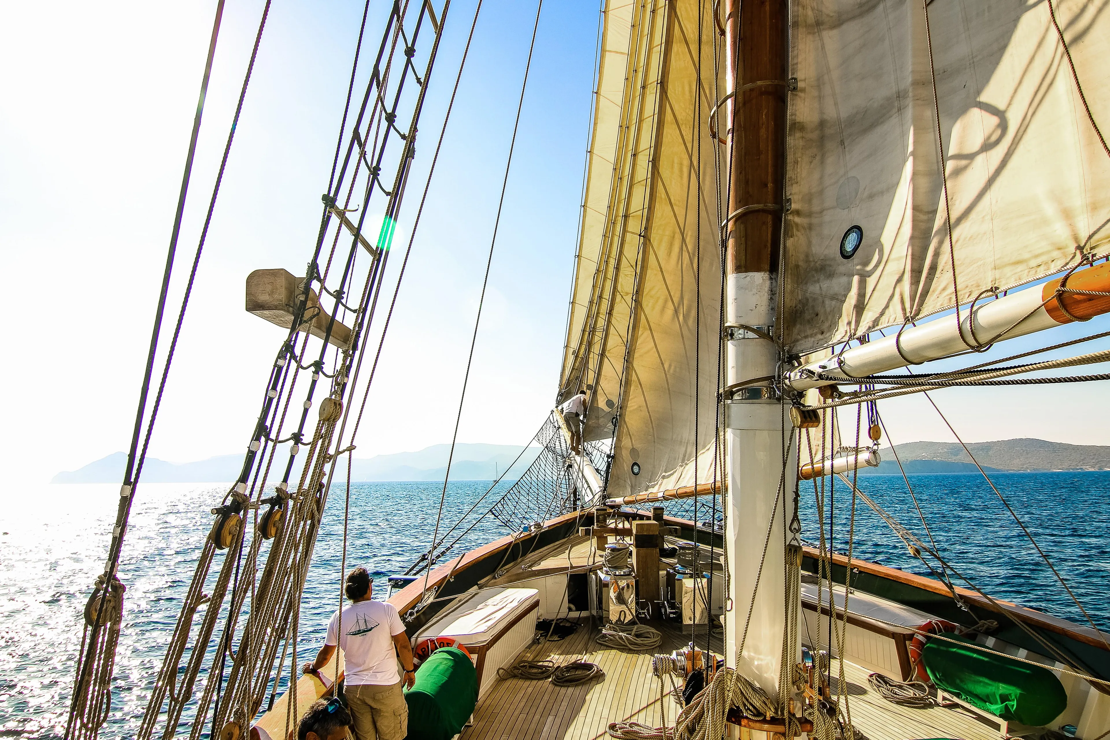 Sailing ship on open sea with crew member, bright sunlight, and distant islands.