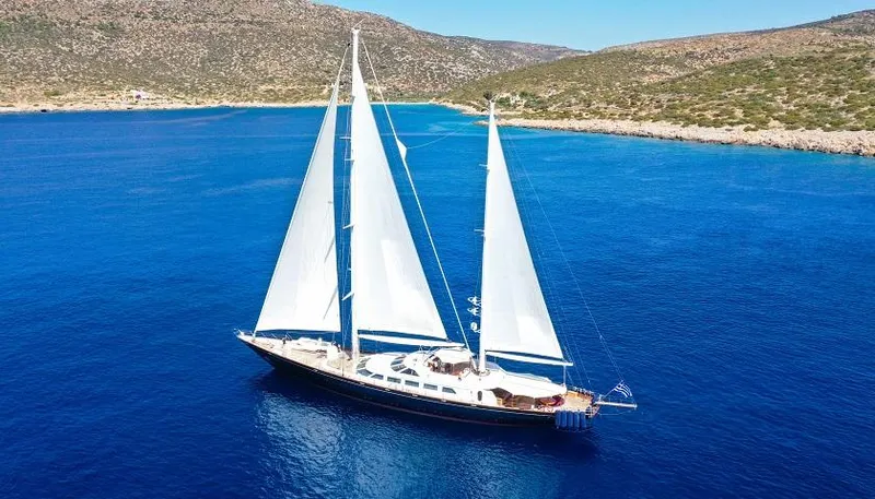 Tamarita Yacht Photos Pics Sailing yacht Perini Navi 46, 1992 model, cruising in clear blue waters near rocky coastline.