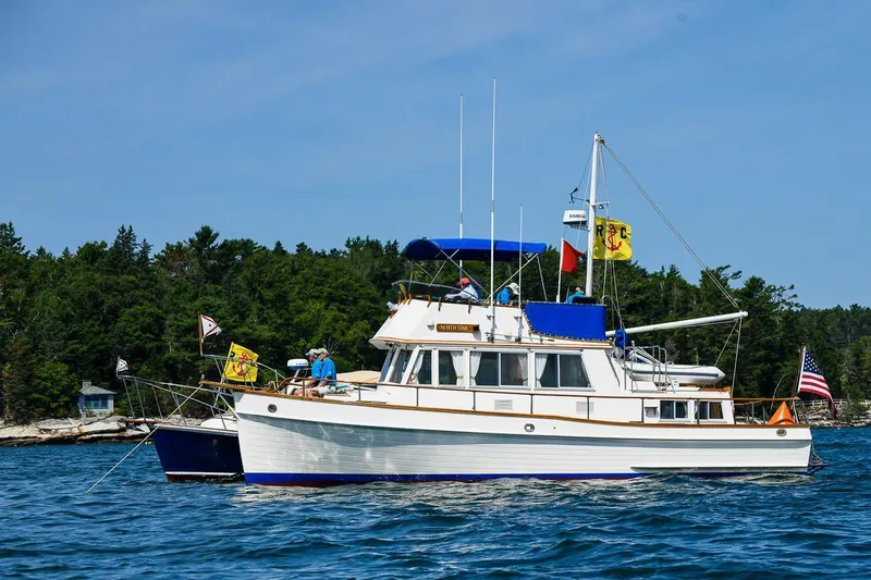 North Star Yacht Photos Pics 1984 Grand Banks Trawler on water, flags flying, surrounded by trees and clear sky.