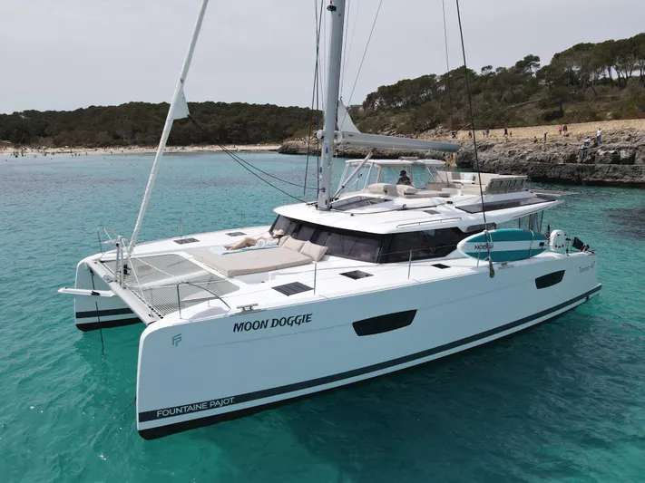 Moon Doggie Yacht Photos Pics 2022 Fountaine Pajot Tanna 47 catamaran sailing in clear turquoise waters near a sandy beach.