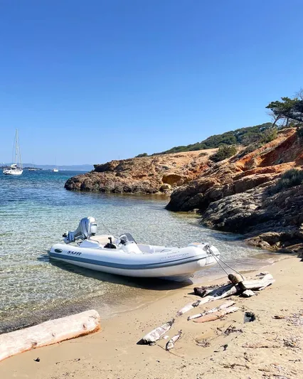 Moon Doggie Yacht Photos Pics Inflatable boat on sandy beach with rocky coastline, clear water, and distant sailboats under blue sky.
