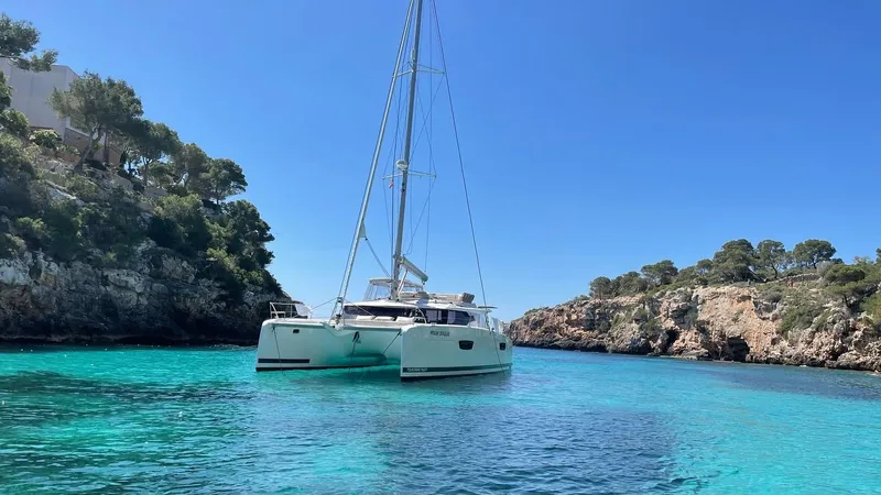 Moon Doggie Yacht Photos Pics 2022 Fountaine Pajot Tanna 47 catamaran in clear turquoise waters, surrounded by rocky cliffs.