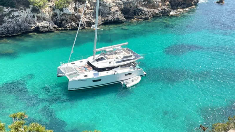 Moon Doggie Yacht Photos Pics 2022 Fountaine Pajot Tanna 47 catamaran in clear turquoise waters near rocky coastline.