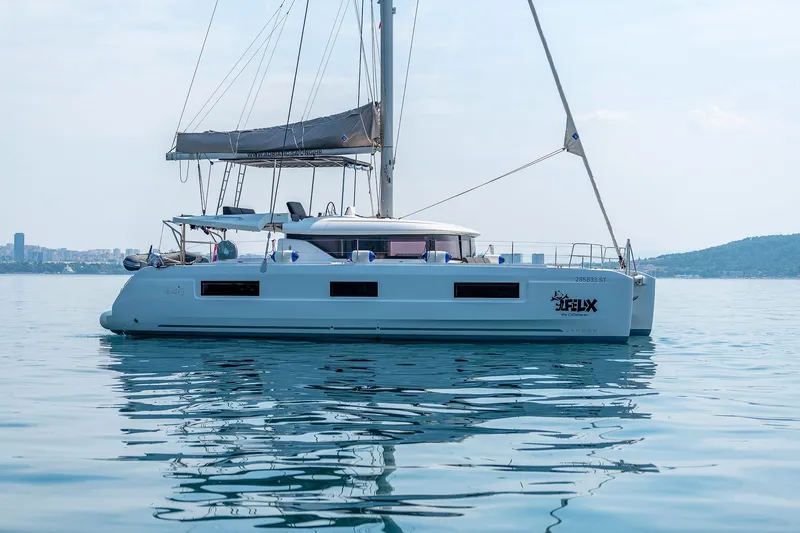  Yacht Photos Pics 2024 Lagoon 450 F catamaran sailing on calm waters, cityscape in background.