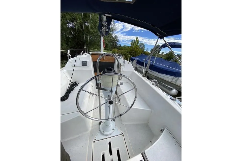 1990 Hunter 27 sailboat cockpit with steering wheel, docked near trees and blue sky.