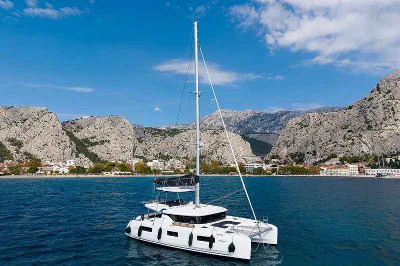 Almissa Yacht Photos Pics 2023 Lagoon 46 catamaran sailing near rocky coastline under clear blue sky.