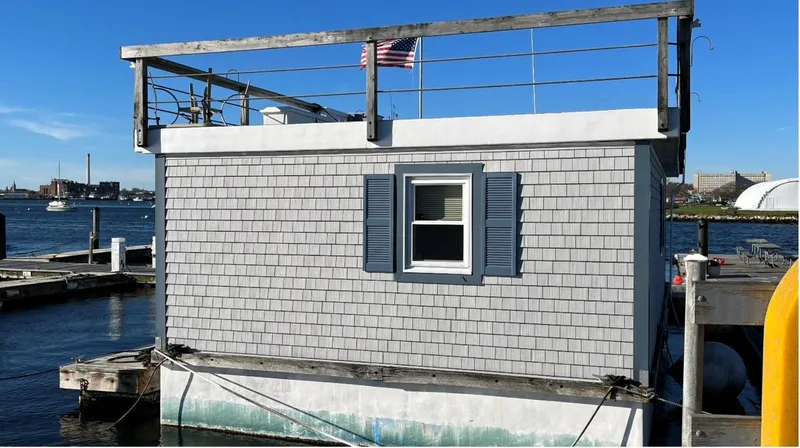  Yacht Photos Pics Custom 1990 houseboat with gray siding, docked by the waterfront, featuring a small window and rooftop railing.